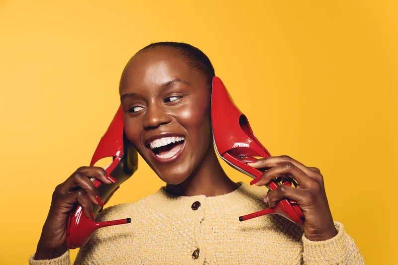 Happy African woman with dark skin holding red high heels near her ears, smiling in front of yellow background. Concept of joy, fashion, and beauty with vibrant colors.