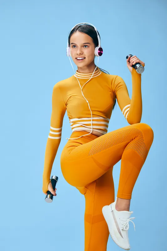 Active woman in bright orange fitness outfit lifts dumbbells and wears headphones in a studio with a blue background, showcasing dynamic workout, balance, and motivation for wellness and healthy