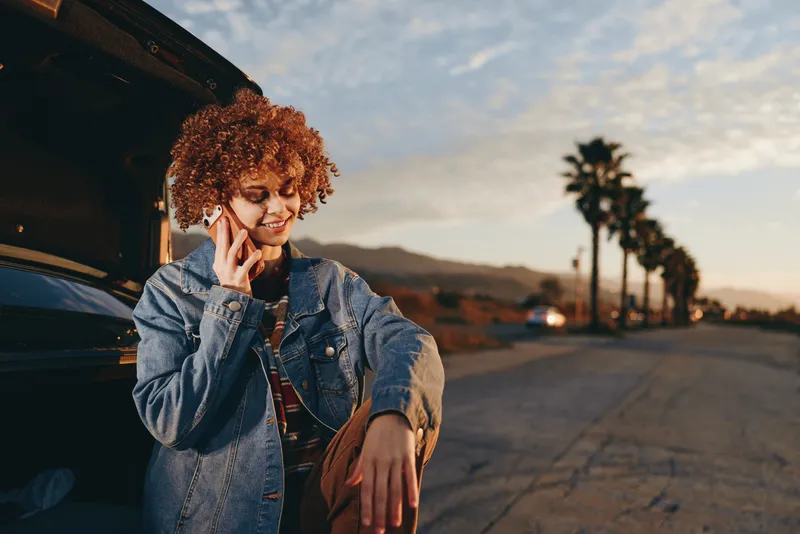 Woman lifestyle at sunset street, woman in denim jacket calling on phone, relaxed smile near parked car, palm trees, warm evening light outdoors, casual fashion portrait by road