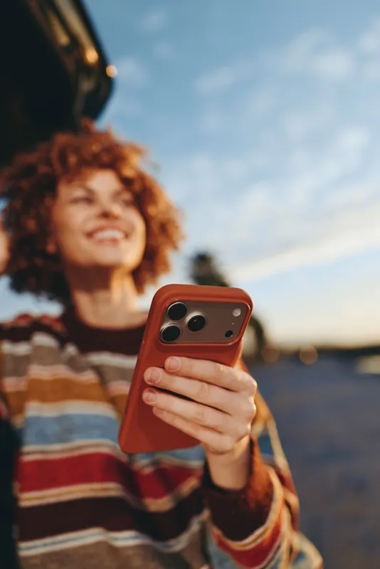 Woman lifestyle outdoors portrait, curly hair smile, holding smartphone with both hands, warm sunset light, casual striped sweater, soft background blur, relaxed confident mood