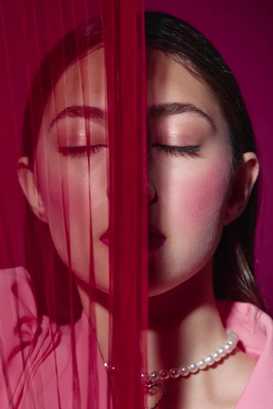 Close-up of young woman with glossy pink makeup and lively cheeks behind sheer red veil. Fashion beauty portrait with pearls necklace and creative lighting concept.