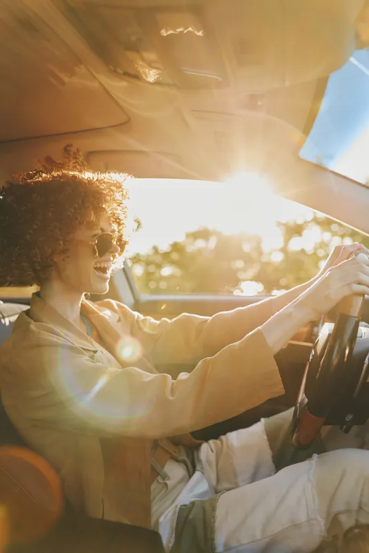 Smiling woman driving a car in warm retro film tones, sunny road scene, casual lifestyle moment, relaxed mood, cheerful energy and nostalgic vibe.