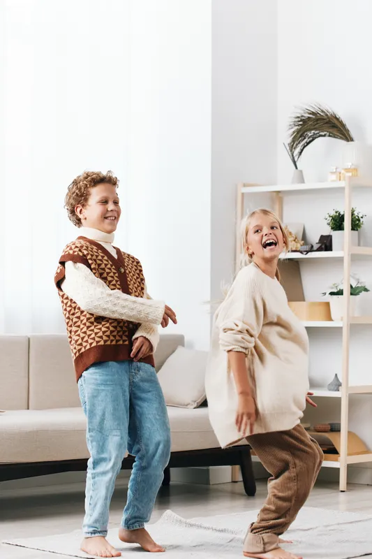 Authentic scene of children at play in a bright living room, capturing spontaneous movement, simple school inspired dress, cheerful genuine energy, and playful drama in a cozy home setting.