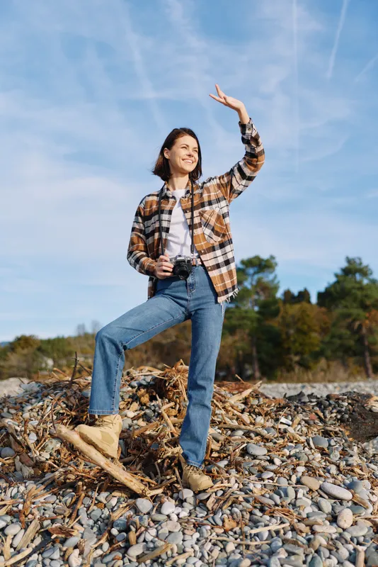 Woman standing on a rocky shoreline in casual outdoor fashion, plaid shirt over a white tee, denim jeans and boots, posing with one arm raised against a bright blue sky.