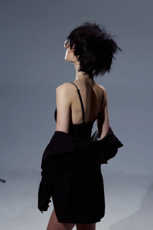 woman in black dress with short hair posing with back turned against gray background