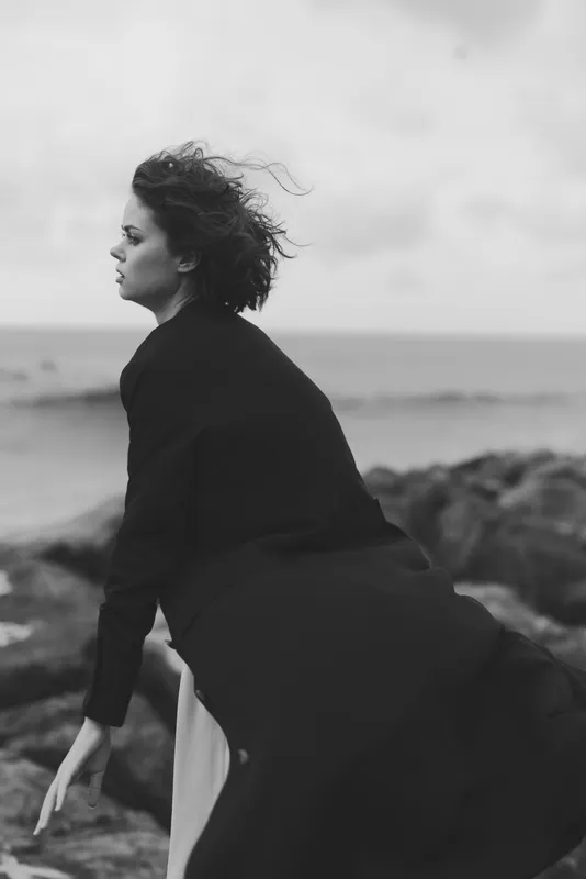 Wind sweeps a figure along a rocky shore in a dark coat, black and white tones, capturing motion and solitude by the sea, dramatic light and minimalist mood.