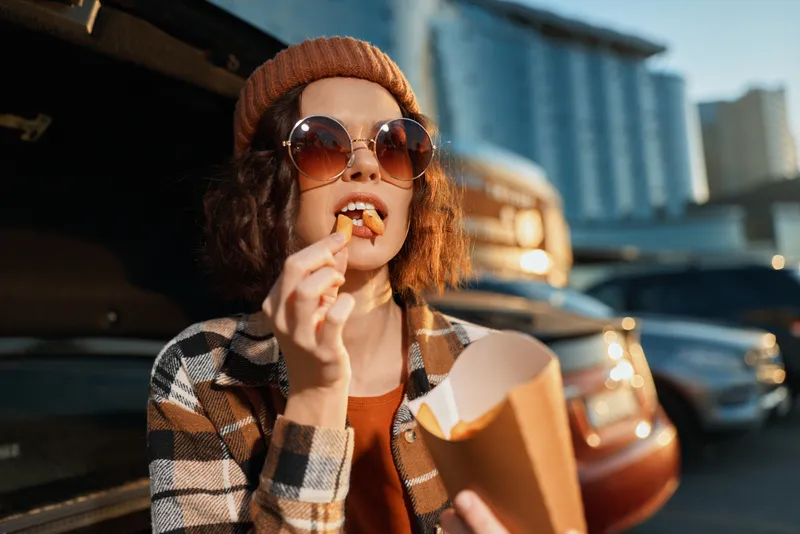 Woman eating fries in parking by car, wearing sunglasses and beanie, casual snack moment with urban vibe, candid authenticity, golden hour glow and emotional storytelling mood.