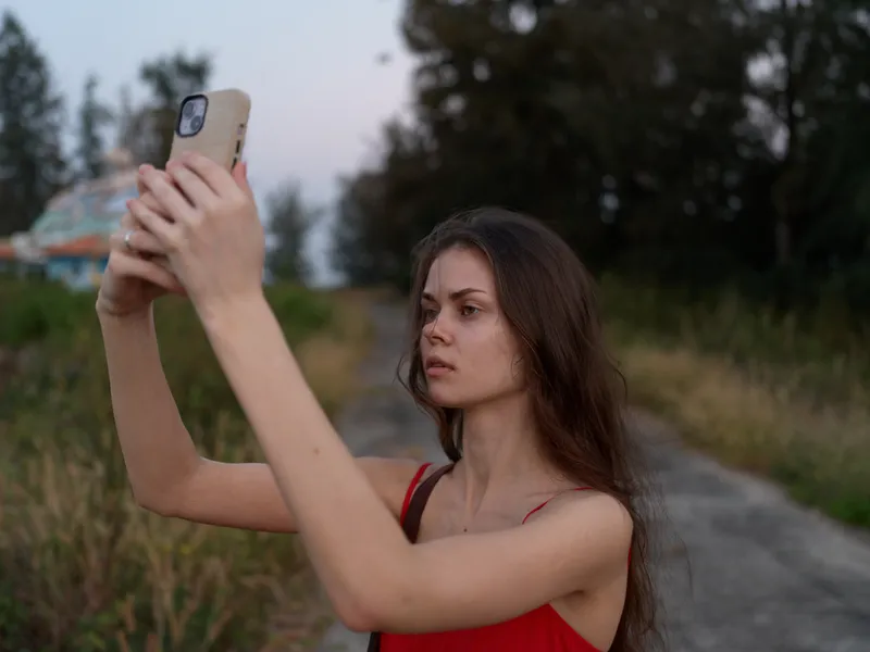 young woman in red dress taking selfie with smartphone outdoors, natural light during golden hour, focused expression, rural path and trees in background