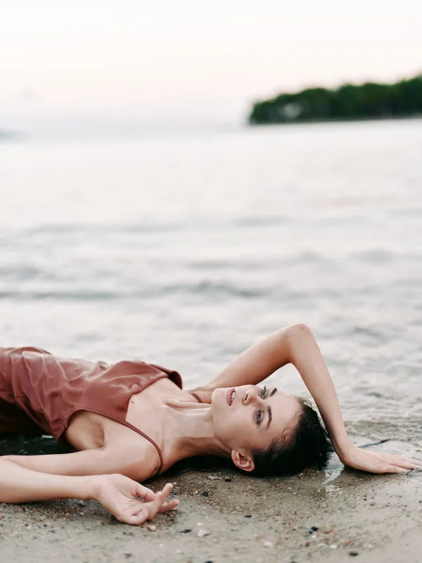 A woman lies on a sandy shore by calm water, wearing a rust dress, one arm raised, eyes closed, embodying a serene moment of relaxation and coastal breeze.