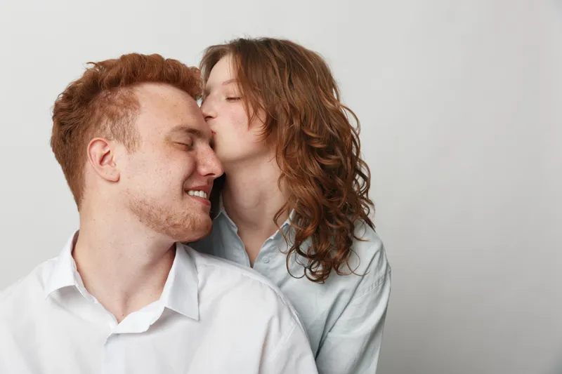 Redhead woman kisses smiling man on cheek in white shirts against plain background. Intimate moment of affection, love, and happiness between young couple with natural expressions.