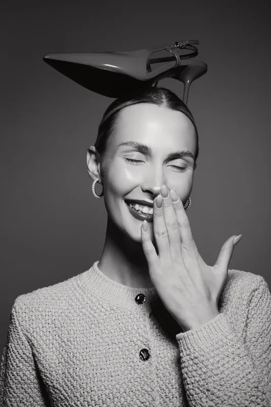 Smiling Caucasian woman with light skin laughs covering mouth. She wears a textured jacket and balances a high heel shoe on her head. Studio portrait with playful mood concept.