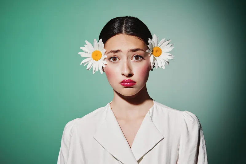 Young woman with fresh natural makeup and bright flowers in her dark hair looks curious on a mint background. Creative beauty fashion portrait emphasizing bold color contrast.