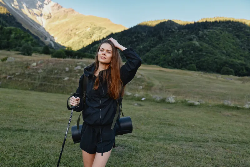 A confident hiker poses in a wide meadow beneath blue skies, holding a trekking pole and smiling at the camera. She wears a dark windbreaker and shorts, embracing outdoor adventure.