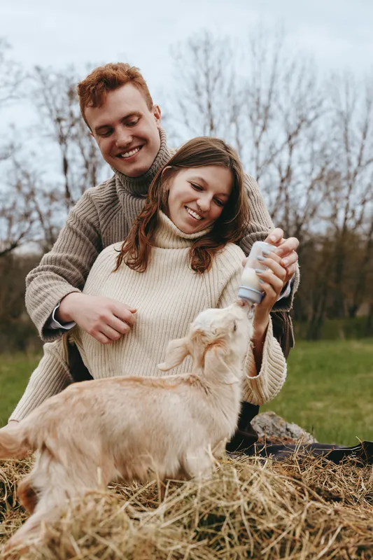 Young couple in cozy sweaters happily bottle-feeds a baby goat outdoors in a natural setting during autumn, enjoying a warm and playful lifestyle moment together.