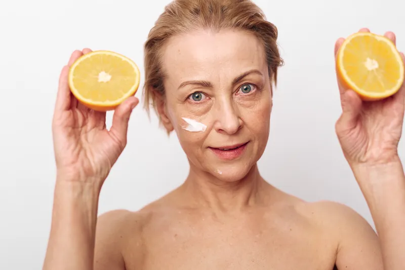 Mature woman with natural skin holds two halves of fresh orange near face with cream on cheek. Concept of healthy skincare, natural beauty, vitamin c, and self-care routine for glowing skin.