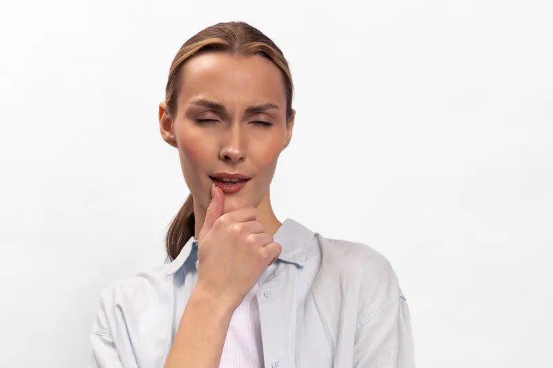 Young woman with closed eyes and thoughtful expression touches chin with hand. Studio portrait on white background. Concept of thinking, decision making, and contemplation.