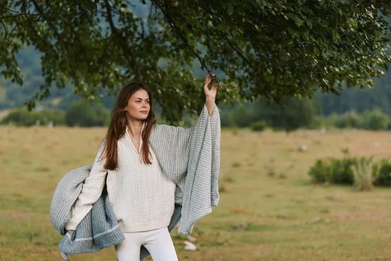 Stylish woman enjoying outdoor life in a field under a leafy tree, wearing a cozy sweater and draped coat. Natural fashion, nature, and relaxed mood.