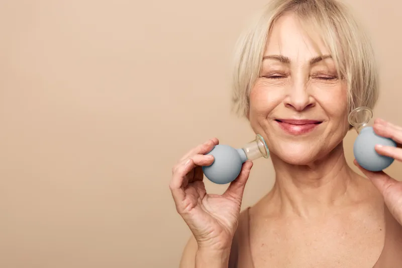 Smiling mature woman with closed eyes holds facial cupping therapy tools near face on beige background. Concept of skincare, wellness, anti aging and natural beauty treatment at home.