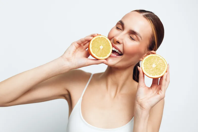 Young woman with long brunette hair in a white tank top holds orange halves near her face and smiles against a light, neutral background, conveying freshness, vitality and a playful healthy vibe.