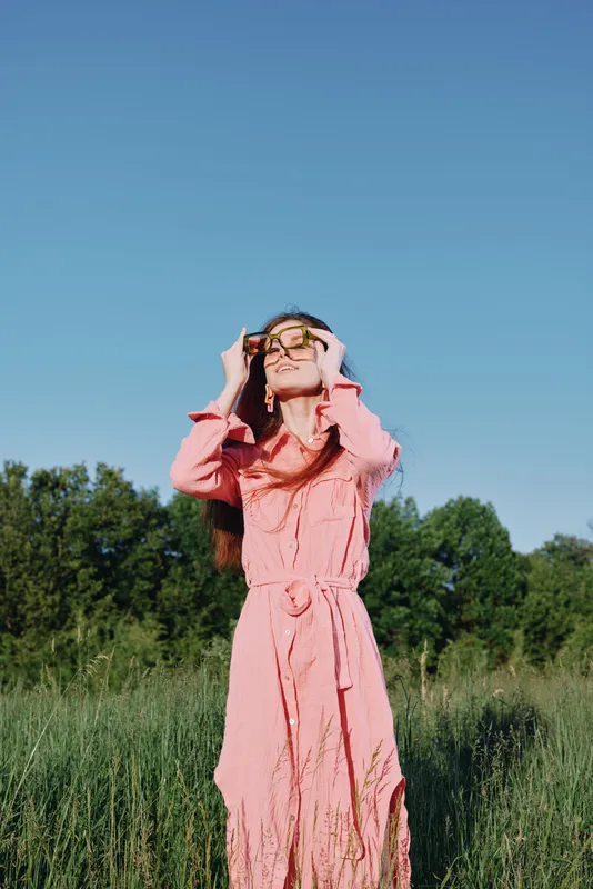 Young woman in a pink dress enjoying a sunny day in a green field, with a relaxed expression and sunglasses, perfect for seasonal fashion and lifestyle concepts