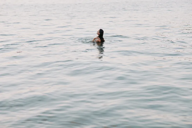 A lone swimmer floats in calm coastal waters, body partly submerged, light ripples around, capturing solitude and peaceful moment beneath a pastel sky.