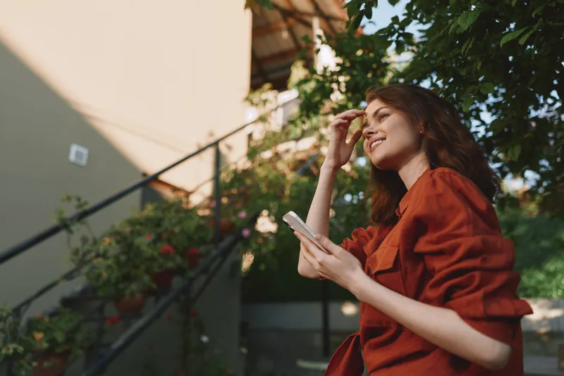 Cheerful woman outdoors, smiling with phone in hand, warm sunlight, relaxed pose, natural colors, leafy backdrop, casual fashion, sunlit moment