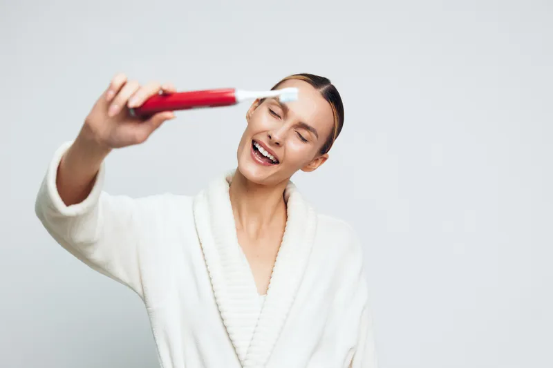 Young woman in a white bathrobe applies white face cream with a brush while smiling with closed eyes against a plain light background.