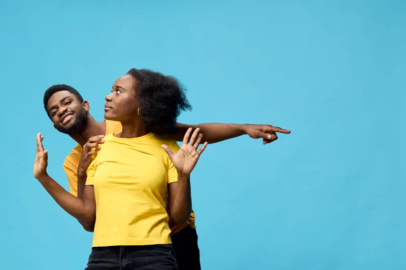 Happy diverse couple posing against a blue background, showcasing playful interaction and vibrant yellow outfits, perfect for joy and diversity themes