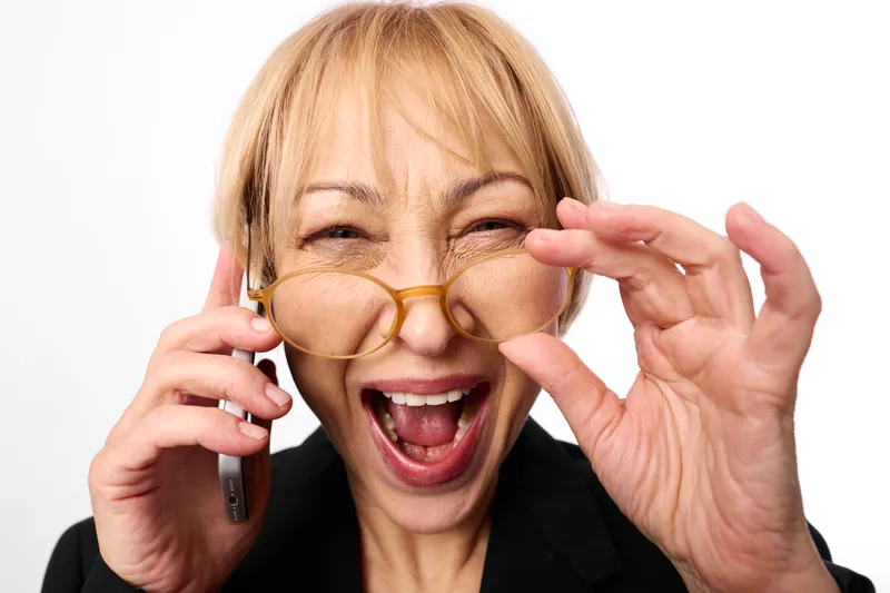 Senior woman with glasses laughing in studio closeup, adjusting gold eyeglasses with both hands, playful surprised expression, white background, soft light