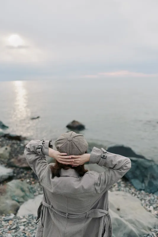 A person in a textured coat and knitted hat stands on rocky shore looking at calm sea under cloudy sky. Concept of solitude, nature, peaceful moment, and cold weather outdoors.