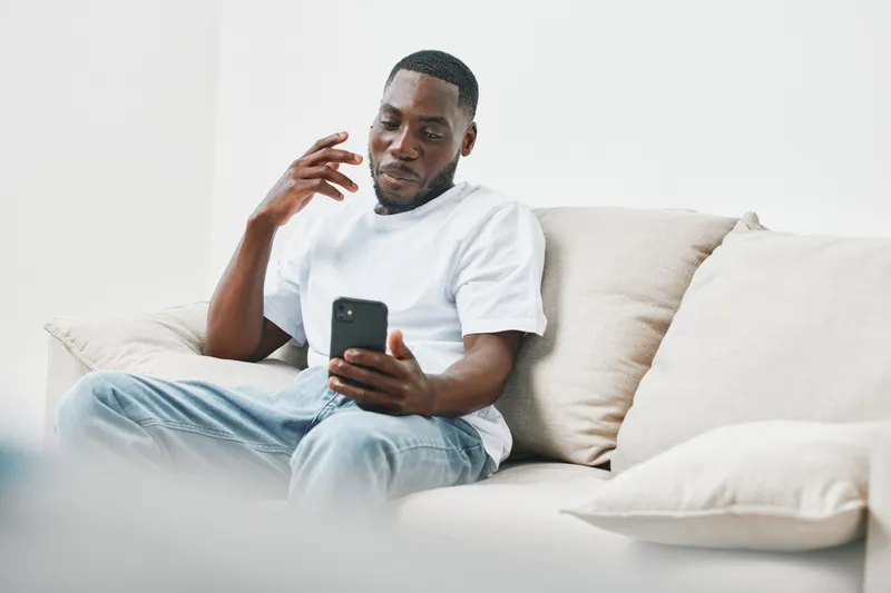 Young Black man wearing a white t shirt and jeans, relaxed on a couch while using a smartphone in a bright, minimalistic room