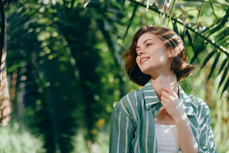 Portrait of a young woman outdoors, basking in sunlight amid lush greenery, wearing a striped green jacket and white top.