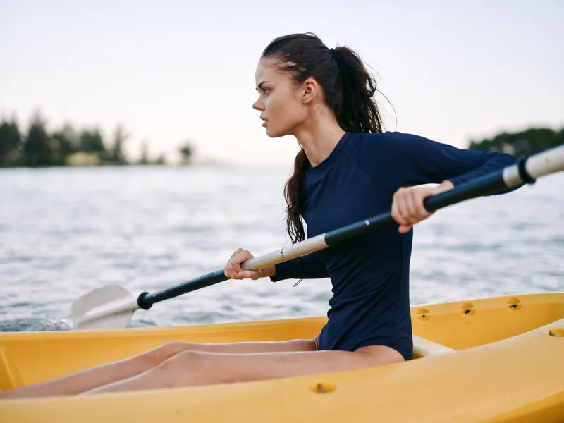 Female rower in a navy athletic suit paddles a bright yellow kayak, concentrating on the stroke as calm water and distant trees create a serene outdoor scene.