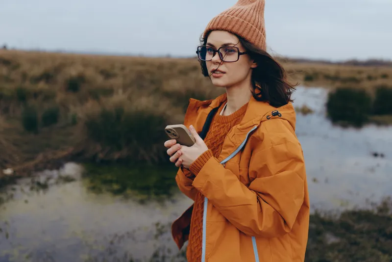 Woman lifestyle outdoors, orange jacket, knit beanie and glasses, holding smartphone, walking with backpack by river, autumn nature landscape, candid travel moment