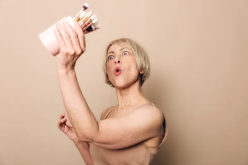 Mature woman with short hair holds a cup full of makeup brushes, looking surprised. Neutral background emphasizes beauty tools and self-care concept for middle aged skin.