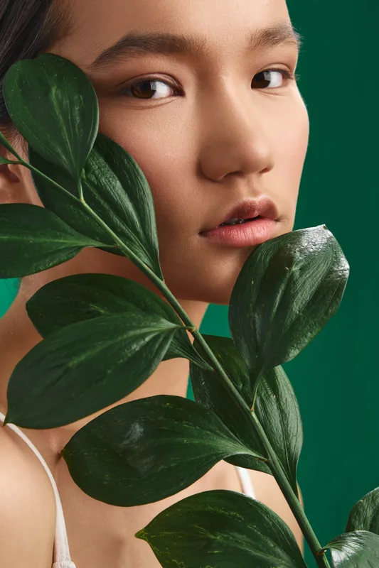 Asian woman gazes softly at the camera as lush green leaves frame her face, creating a serene beauty portrait with natural textures and gentle lighting.