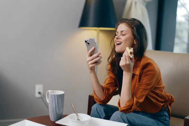 Весёлая молодая женщина держит в одной руке печенье, а в другой — смартфон, сидя в стильной гостиной с тёплой атмосферой, что подчёркивает комфорт и современность