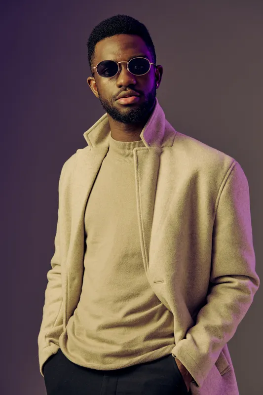 Stylish young man with sunglasses in a beige coat and light sweater, exuding confidence against a soft purple background, perfect for fashion and lifestyle themes