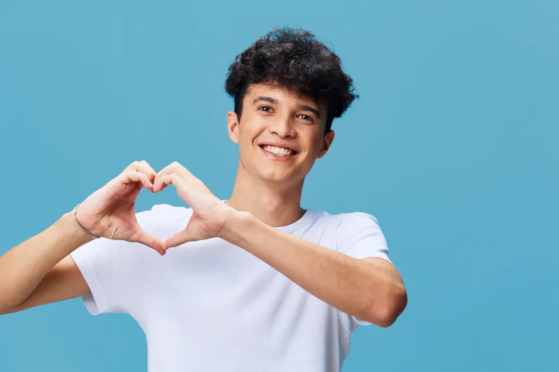 Smiling man making heart shape with hands, curly hair, wearing white t-shirt, light blue background, cheerful expression, casual style, young adult male.