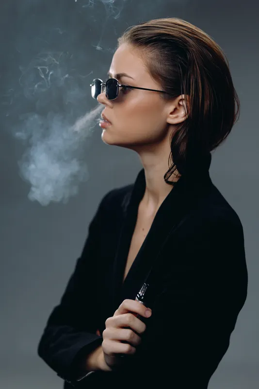 Young woman vaping with sunglasses exhaling smoke wearing black outfit on dark background