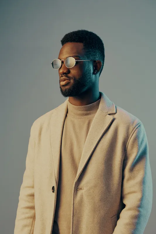 Stylish young man in a light coat and round glasses against a pastel background, exuding confidence and modernity in a seasonal fashion shoot