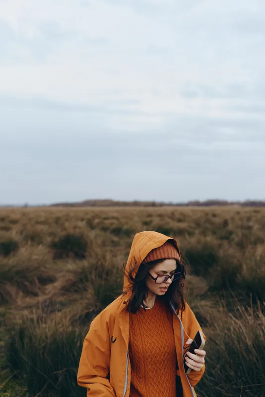 Woman in orange hooded coat in grassy field, outdoors lifestyle, autumn nature, she looks at phone while walking, cloudy sky behind, calm leisure mood