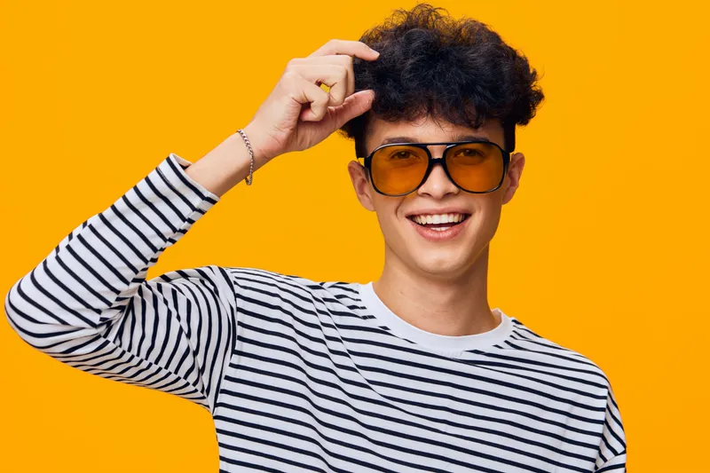 smiling man wearing striped shirt and sunglasses posing against bright orange background with curly hair and joyful expression, casual style portrait