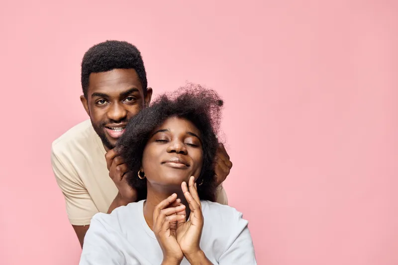 A joyful couple with natural hairstyles poses together against a pastel pink background, showcasing love, happiness, and connection in a stylish manner