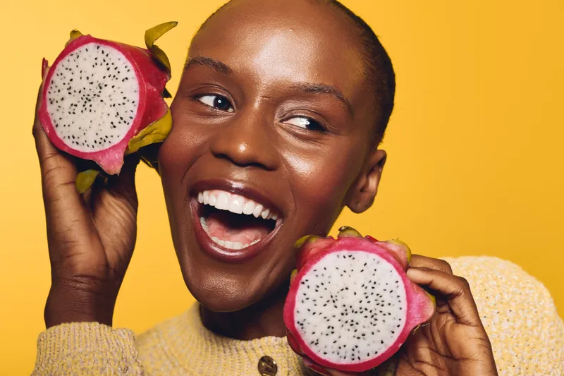 Happy smiling African woman with dark skin holds two halves of ripe dragon fruit near her face on yellow background. Concept of healthy lifestyle, tropical fruit, and natural beauty.