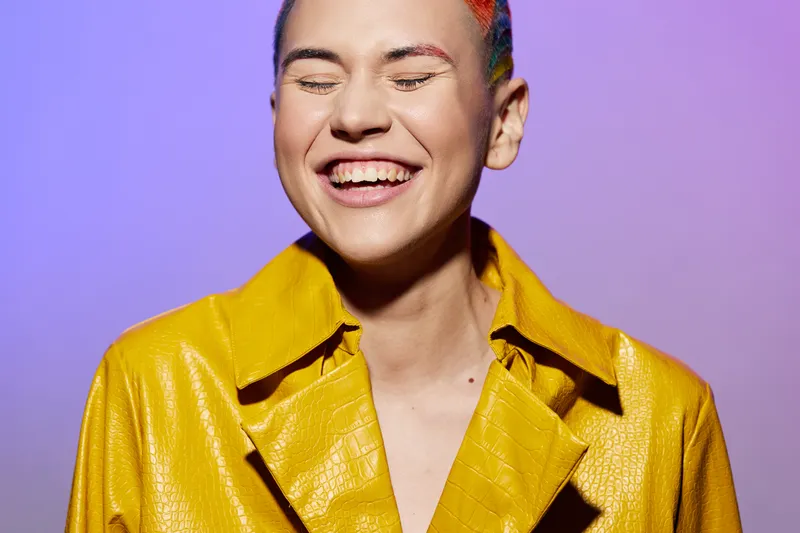 Young woman smiling joyfully with eyes closed wearing a vibrant yellow coat against a soft purple background, showcasing carefree and colorful fashion style.