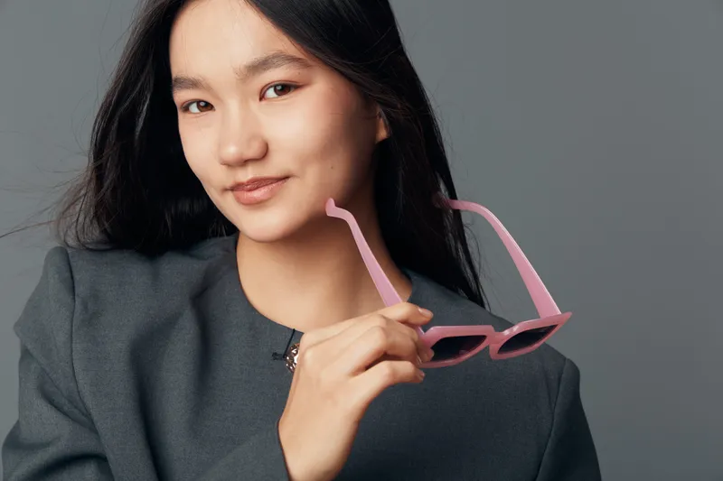 Young woman with long hair holding pink sunglasses close to her face with a slight smile wearing a gray jacket against neutral background in studio portrait photography.