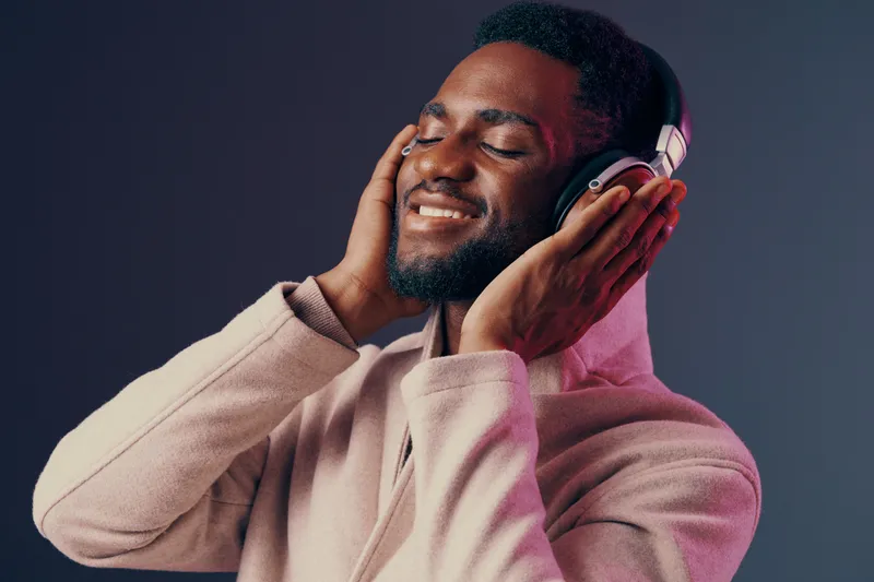 Young African man enjoying music with headphones, smiling happily in a stylish outfit, against a colorful background, conveying relaxation and joy