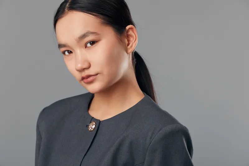 Young woman with long dark hair in ponytail wearing dark gray formal jacket looking confidently at camera with calm expression on gray background in studio portrait.