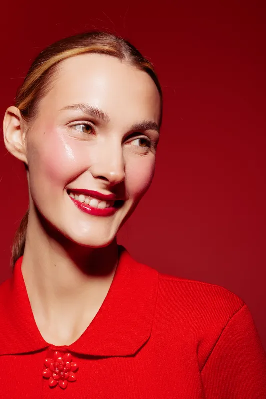 Smiling woman with shiny skin and red lipstick looks to the side wearing a red top against a red background. Concept of beauty, makeup, fashion, and confident expression.